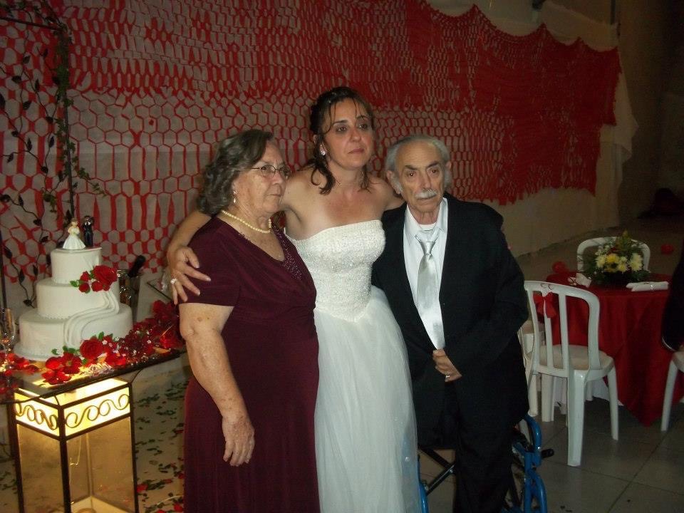 Uma noiva posa para foto com seus pais vestidos para o casamento do lado de um bolo branco.
